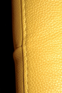 A close-up view of a textured yellow leather surface, displaying visible stitching along a seam. The background is dark, enhancing the texture and color of the leather.