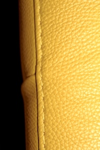A close-up view of a textured yellow leather surface, displaying visible stitching along a seam. The background is dark, enhancing the texture and color of the leather.