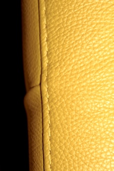 A close-up view of a textured yellow leather surface, displaying visible stitching along a seam. The background is dark, enhancing the texture and color of the leather.