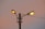 A pair of bright street lamps are mounted on a single pole, with each lamp illuminated casting a warm glow. The background sky displays a gradient of soft pink and light blue hues, suggesting either dawn or dusk. Electrical wires are visible, connecting the lamps to a power source.