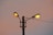 A pair of bright street lamps are mounted on a single pole, with each lamp illuminated casting a warm glow. The background sky displays a gradient of soft pink and light blue hues, suggesting either dawn or dusk. Electrical wires are visible, connecting the lamps to a power source.