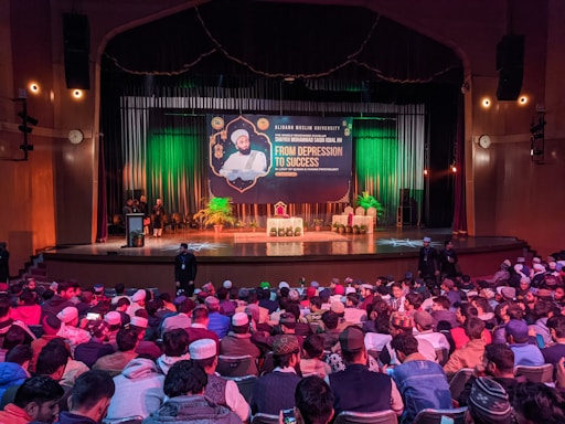 A large audience is gathered in an auditorium for a lecture. The stage features a banner with the text 'From Depression to Success' and the speaker's details. The audience appears attentive, with most people seated. There are potted plants on the stage, and the room is lit with warm lighting.