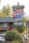 A colorful motel sign with a crown design advertising the Crown Motel with private beach access and no vacancy. Below it is another sign indicating Lakefront Lodging. The background features a building with a balcony, surrounded by trees and parked vehicles. The environment suggests a serene, natural setting.