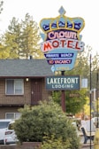 A colorful motel sign with a crown design advertising the Crown Motel with private beach access and no vacancy. Below it is another sign indicating Lakefront Lodging. The background features a building with a balcony, surrounded by trees and parked vehicles. The environment suggests a serene, natural setting.
