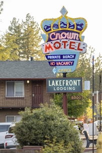 A colorful motel sign with a crown design advertising the Crown Motel with private beach access and no vacancy. Below it is another sign indicating Lakefront Lodging. The background features a building with a balcony, surrounded by trees and parked vehicles. The environment suggests a serene, natural setting.