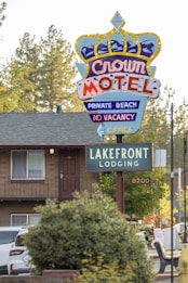 A colorful motel sign with a crown design advertising the Crown Motel with private beach access and no vacancy. Below it is another sign indicating Lakefront Lodging. The background features a building with a balcony, surrounded by trees and parked vehicles. The environment suggests a serene, natural setting.