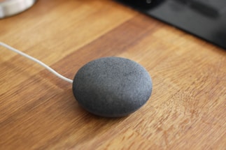 A small, round, gray smart speaker with a fabric covering is placed on a wooden surface. A white cable is attached to the speaker, indicating it is plugged in.