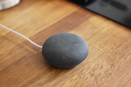 A small, round, gray smart speaker with a fabric covering is placed on a wooden surface. A white cable is attached to the speaker, indicating it is plugged in.