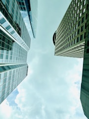Tall skyscrapers with reflective glass facades are viewed from below, converging towards the center of the image against a backdrop of a cloudy sky.