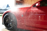 A red car is being washed with water spraying over its body. The vehicle's front side, including a flared wheel arch and front wheel with black rims, is visible. Water droplets and mist obscure part of the image, reflecting sunlight. In the background, there is a blurred blue vehicle and a wooden panel wall inside a garage or car wash.