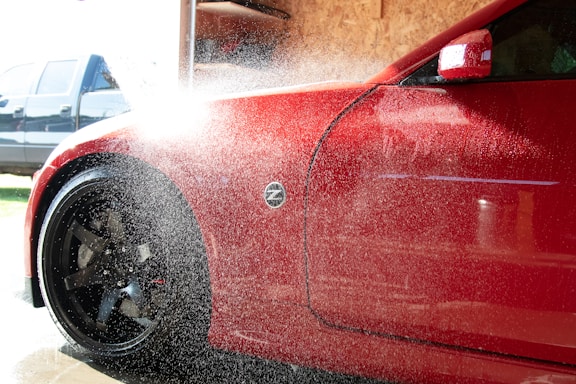 A red car is being washed with water spraying over its body. The vehicle's front side, including a flared wheel arch and front wheel with black rims, is visible. Water droplets and mist obscure part of the image, reflecting sunlight. In the background, there is a blurred blue vehicle and a wooden panel wall inside a garage or car wash.