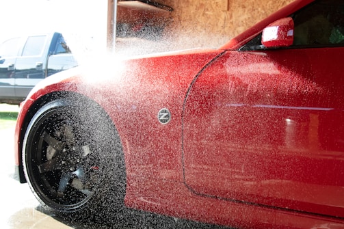 A red car is being washed with water spraying over its body. The vehicle's front side, including a flared wheel arch and front wheel with black rims, is visible. Water droplets and mist obscure part of the image, reflecting sunlight. In the background, there is a blurred blue vehicle and a wooden panel wall inside a garage or car wash.