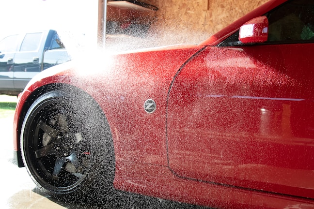 A red car is being washed with water spraying over its body. The vehicle's front side, including a flared wheel arch and front wheel with black rims, is visible. Water droplets and mist obscure part of the image, reflecting sunlight. In the background, there is a blurred blue vehicle and a wooden panel wall inside a garage or car wash.