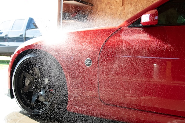 A red car is being washed with water spraying over its body. The vehicle's front side, including a flared wheel arch and front wheel with black rims, is visible. Water droplets and mist obscure part of the image, reflecting sunlight. In the background, there is a blurred blue vehicle and a wooden panel wall inside a garage or car wash.