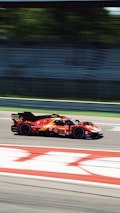 A red race car speeds along a track, with motion blur conveying high velocity. The background features green foliage behind a mostly empty grandstand, suggesting a professional racing environment.