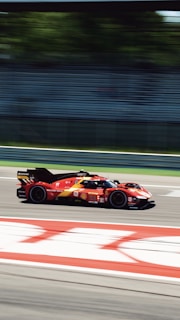 A red race car speeds along a track, with motion blur conveying high velocity. The background features green foliage behind a mostly empty grandstand, suggesting a professional racing environment.