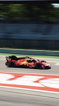 A red race car speeds along a track, with motion blur conveying high velocity. The background features green foliage behind a mostly empty grandstand, suggesting a professional racing environment.