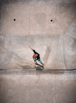 A person wearing a red helmet and green pants is performing a skateboarding trick in a concrete skate park. The setting appears to be an empty area with smooth, curved surfaces designed for skating. Shadows fall across the surface, and the person seems to be captured mid-action.