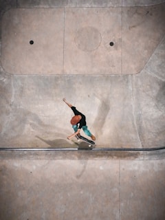 A person wearing a red helmet and green pants is performing a skateboarding trick in a concrete skate park. The setting appears to be an empty area with smooth, curved surfaces designed for skating. Shadows fall across the surface, and the person seems to be captured mid-action.