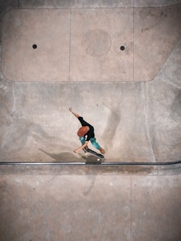 A person wearing a red helmet and green pants is performing a skateboarding trick in a concrete skate park. The setting appears to be an empty area with smooth, curved surfaces designed for skating. Shadows fall across the surface, and the person seems to be captured mid-action.
