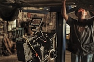 A professional film camera is set up in a workshop or garage-like setting. The background is filled with tools and machinery, indicating a working environment. A person is partially seen, engaged in some task, adding to the industrial feel of the image.