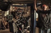 A professional film camera is set up in a workshop or garage-like setting. The background is filled with tools and machinery, indicating a working environment. A person is partially seen, engaged in some task, adding to the industrial feel of the image.