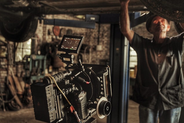 A professional film camera is set up in a workshop or garage-like setting. The background is filled with tools and machinery, indicating a working environment. A person is partially seen, engaged in some task, adding to the industrial feel of the image.