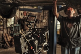 A professional film camera is set up in a workshop or garage-like setting. The background is filled with tools and machinery, indicating a working environment. A person is partially seen, engaged in some task, adding to the industrial feel of the image.