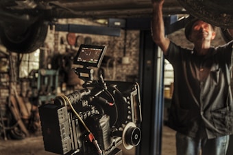 A professional film camera is set up in a workshop or garage-like setting. The background is filled with tools and machinery, indicating a working environment. A person is partially seen, engaged in some task, adding to the industrial feel of the image.