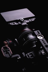 A professional camera setup with a mounted lens, accompanied by various metal components and attachments, against a dark black background. The lens features detailed markings and the setup includes a matte box or filter mounted in front.
