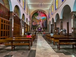 A spacious church interior with high ceilings and rows of wooden pews. Stained glass windows line the upper walls, casting colorful light into the space. A large, vibrant mural depicting a religious scene is featured prominently at the front, above the altar. Several people are seated, engaged in prayer or reflection.