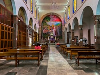A spacious church interior with high ceilings and rows of wooden pews. Stained glass windows line the upper walls, casting colorful light into the space. A large, vibrant mural depicting a religious scene is featured prominently at the front, above the altar. Several people are seated, engaged in prayer or reflection.