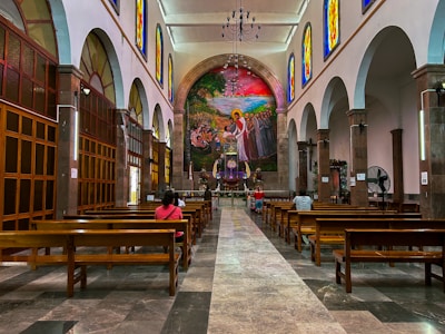 A spacious church interior with high ceilings and rows of wooden pews. Stained glass windows line the upper walls, casting colorful light into the space. A large, vibrant mural depicting a religious scene is featured prominently at the front, above the altar. Several people are seated, engaged in prayer or reflection.