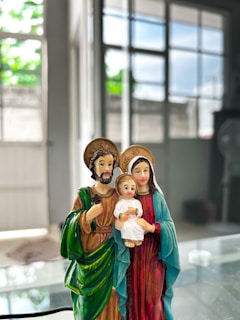 A small, colorful statue depicts a family with a child. The man has a green robe, the woman is dressed in a red and blue outfit, and the child is holding a small object. Behind them is a bright room with large windows, allowing natural light to illuminate the scene.