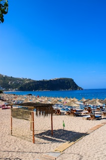 A sunlit sandy beach with multiple rows of straw parasols and deck chairs, overlooking a calm blue sea. In the distance, there is a rocky hill with a few buildings scattered across it. The sky is clear and vibrant, adding to the serene coastal atmosphere.