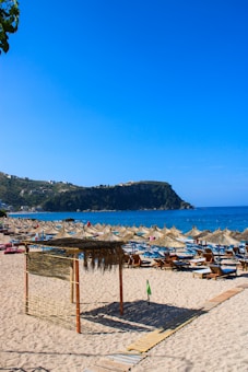 A sunlit sandy beach with multiple rows of straw parasols and deck chairs, overlooking a calm blue sea. In the distance, there is a rocky hill with a few buildings scattered across it. The sky is clear and vibrant, adding to the serene coastal atmosphere.
