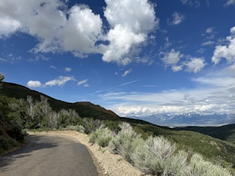 A scenic landscape featuring a winding road through lush green hills. In the background, there are majestic snow-capped mountains under a sky filled with fluffy white clouds.