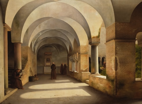 A serene walkway with arched ceilings and columns cast subtle shadows. Monks in robes are scattered around; one sits reading, while two others stand in conversation further down the corridor. Sunlight filters through small windows, illuminating the stone and brick textures. The atmosphere feels calm and introspective.
