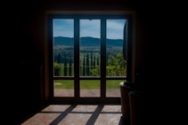 A bright landscape view captured through a large window, showcasing rolling hills and tall cypress trees under a blue sky. The foreground features a cozy interior space with dark shadows cast on a tiled floor.