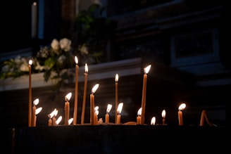 A collection of lit candles of various heights is placed on a dark surface, casting a warm glow in a dimly lit environment. Soft shadows and flickering flames create an intimate and serene atmosphere. In the background, there are blurred white flowers and greenery, adding a touch of softness to the scene.