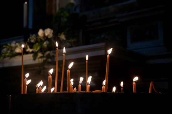 A collection of lit candles of various heights is placed on a dark surface, casting a warm glow in a dimly lit environment. Soft shadows and flickering flames create an intimate and serene atmosphere. In the background, there are blurred white flowers and greenery, adding a touch of softness to the scene.