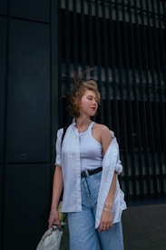A young person stands against a dark-colored background with vertical lines, wearing a white shirt over a tank top and blue jeans. The wind is blowing their hair slightly, and they hold a newspaper. The overall look is casual and relaxed.