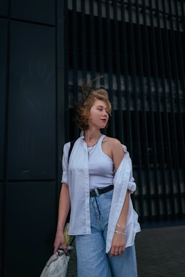 A young person stands against a dark-colored background with vertical lines, wearing a white shirt over a tank top and blue jeans. The wind is blowing their hair slightly, and they hold a newspaper. The overall look is casual and relaxed.