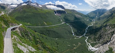 A scenic mountainous landscape with lush green valleys, winding roads, and a distant glacier. A river cuts through the valley, surrounded by rocky terrain and patches of trees. The sky is partly cloudy, providing a bright, natural backdrop.