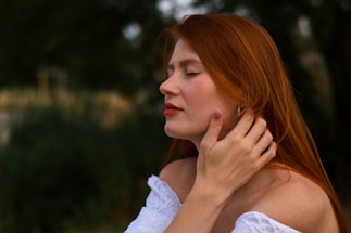 A woman with long red hair is standing outdoors with her eyes closed, gently touching her neck with her hand. She is wearing a white off-shoulder top. The background is blurred, emphasizing the serene expression on her face.