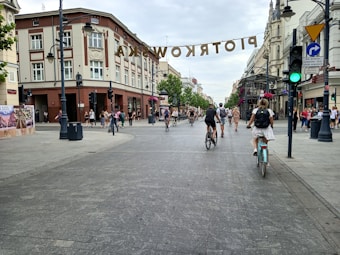A city street scene with people walking and riding bicycles. There are historical buildings on the sides of the street. Hanging above the street is a sign that reads 'PIOTRKOWSKA'. The sky is overcast, and traffic lights and street lamps are visible.