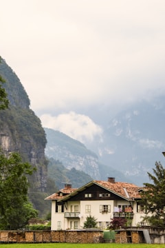 A picturesque white house with a red-tiled roof situated amidst lush greenery, with a backdrop of towering mountains partially shrouded in mist.
