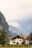 A picturesque white house with a red-tiled roof situated amidst lush greenery, with a backdrop of towering mountains partially shrouded in mist.