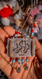 A detailed ornament with intricate patterns and Arabic calligraphy is held in a hand. The ornament is adorned with beads in red, turquoise, and light blue colors, and small hanging charms, including one in the shape of a flower. The background consists of various fluffy keychains in red, white, grey, and pink.