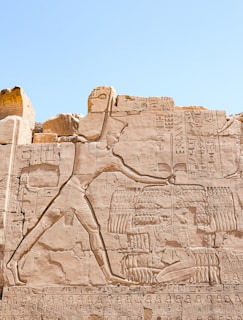 A detailed carving on an ancient stone wall depicting an Egyptian figure adorned with traditional garments and crown, engaged in a ceremonial stance. Hieroglyphics surround the figure, with intricate details of wings and other symbolic elements etched into the stone. The background appears to be part of a historical site with fragments of partially preserved carvings.
